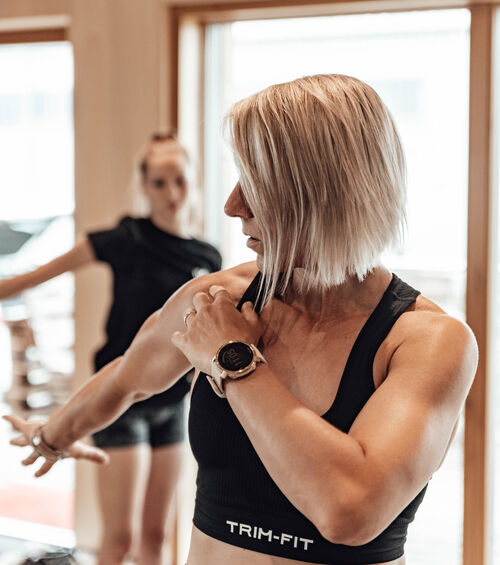 Frau mit kurzen blonden Haaren, die ihren Arm ausstreckt und eine Smartwatch trägt, mit einer weiteren Person, die im Hintergrund verschwommen ist.