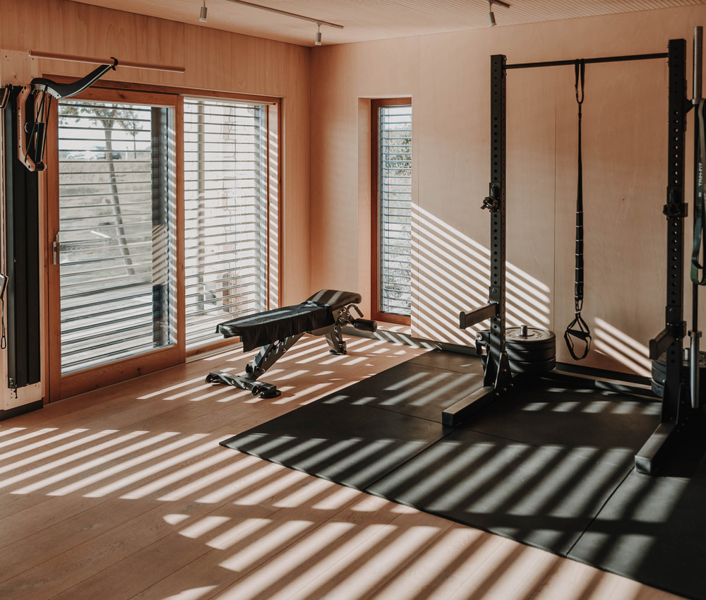 Ein sonnendurchfluteter Fitnessraum mit einer Bank, einem Power Rack und großen Fenstern, die gestreifte Schatten auf den Boden werfen.