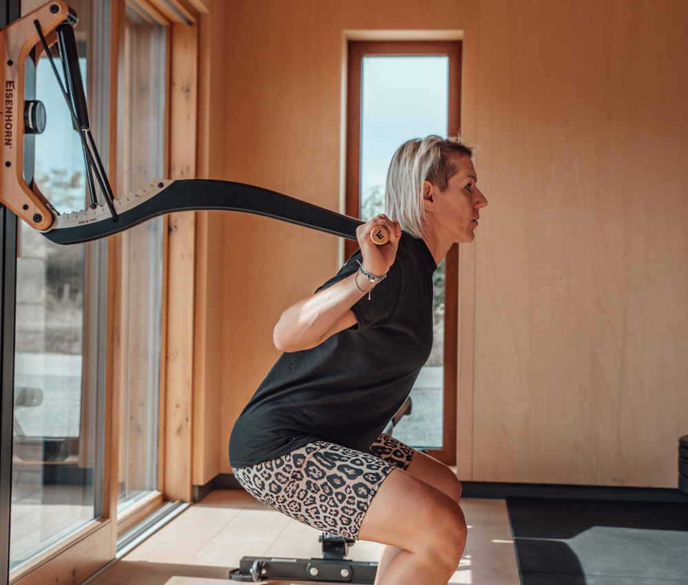 Eine Person mit kurzen blonden Haaren macht Kniebeugen mit einer Langhantel in einem hellen, modernen Fitnessstudio mit Holzwänden.