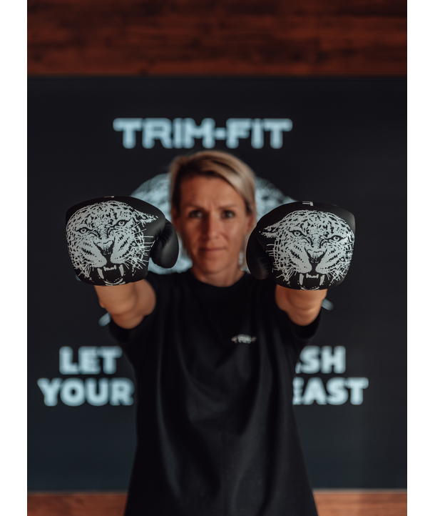 Frau in Boxhandschuhen mit Tigerabdrücken posiert vor einem Trim-Fit-Schild, Arme nach vorne ausgestreckt.