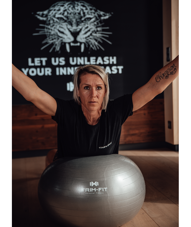 Frau streckt die Arme hinter dem Gymnastikball nach oben, mit Wandmalerei im Fitnessstudio und motivierendem Text im Hintergrund.