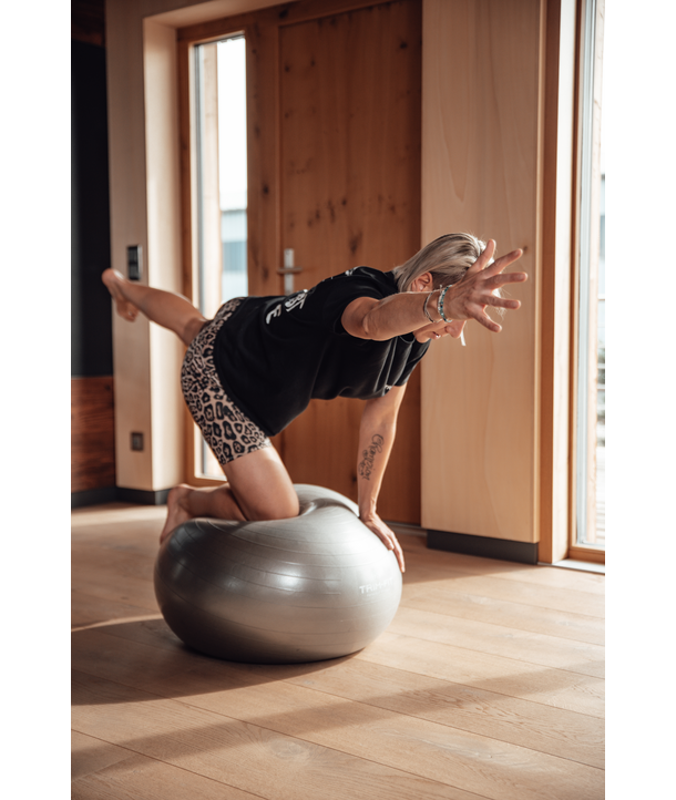 Person, die im Haus auf einem Gymnastikball balanciert, einen Arm ausstreckt und ein Bein anhebt, in schwarzem Hemd und Shorts.