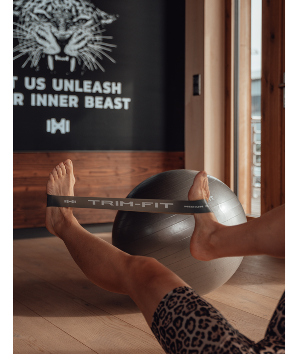 Eine Person dehnt ihre Beine mit einem Widerstandsband in der Nähe eines Gymnastikballs in einem Fitnessstudio mit einem motivierenden Wandschild.