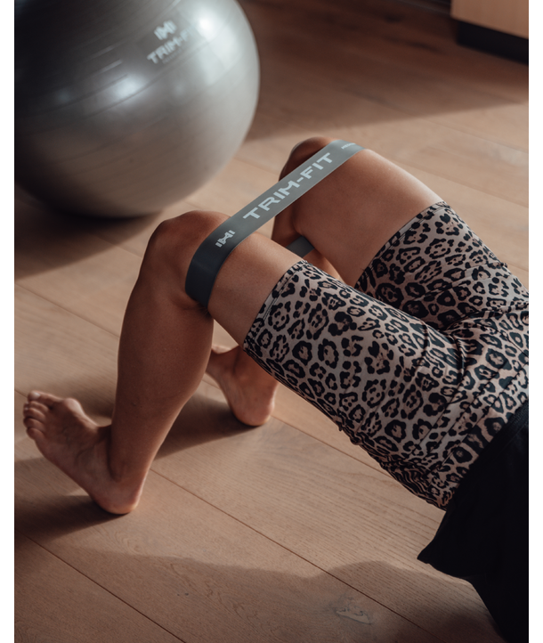 Eine Person trainiert auf einem Holzboden mit einem Widerstandsband über den Knien und einem Gymnastikball in der Nähe.