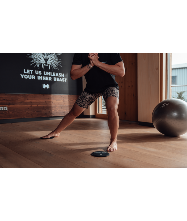 Eine Person macht eine seitliche Ausfallschritt-Übung in einem Raum, trägt Shorts mit Leopardenmuster und steht auf einem Holzboden.