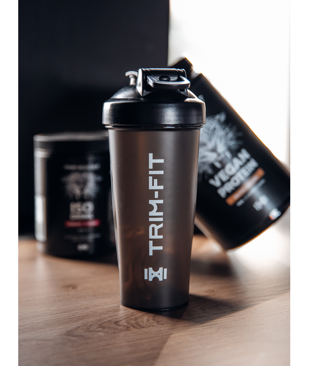 Eine TRIM-FIT-Shakerflasche auf einem Tisch mit Eiweißergänzungsbehältern im Hintergrund.