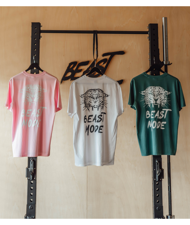 Drei BEAST MODE-T-Shirts in Rosa, Weiß und Grün hängen an einem Turnhallenregal an einer Holzwand.