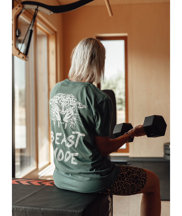 Frau mit blonden Haaren hebt eine Hantel, trägt ein Beast Mode-Shirt und sitzt in einem Fitnessstudio.