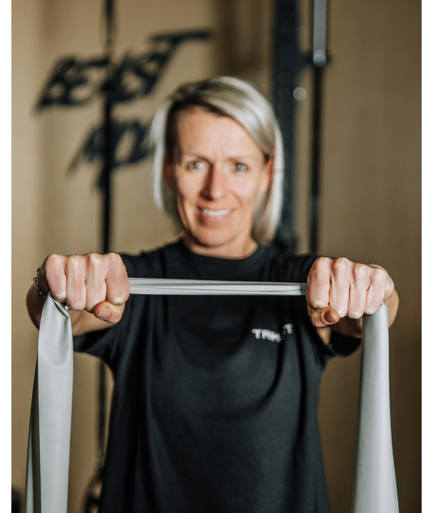 Frau dehnt ein Widerstandsband mit beiden Händen, lächelnd, Beast-Mode-Schild im Hintergrund verschwommen.