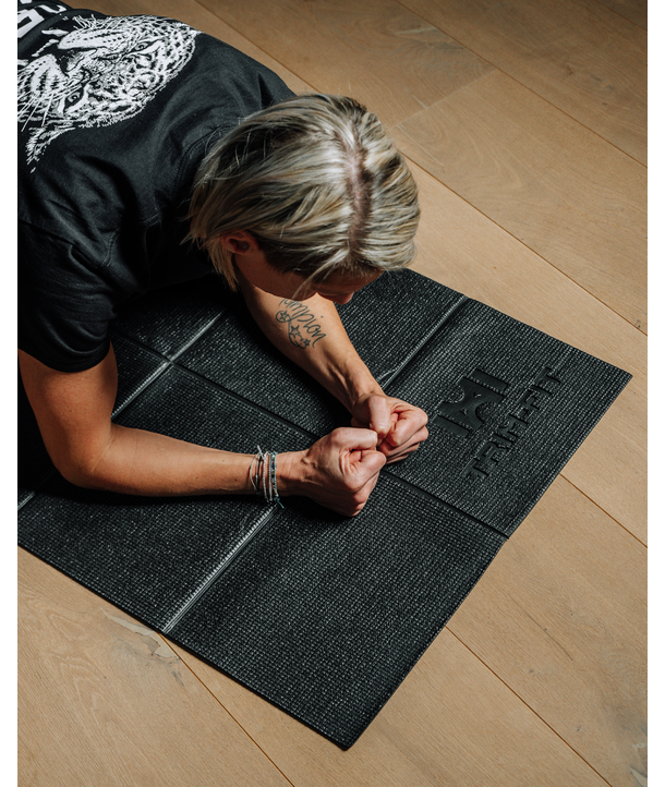 Person mit kurzen blonden Haaren, die eine Planke auf einer schwarzen Yogamatte auf einem Holzboden macht.