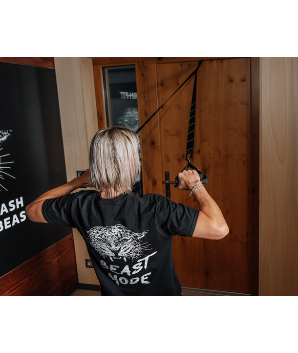 Person mit blondem Haar, die Widerstandsbänder benutzt, die an einer Holztür verankert sind, und ein BEAST MODE-T-Shirt trägt.