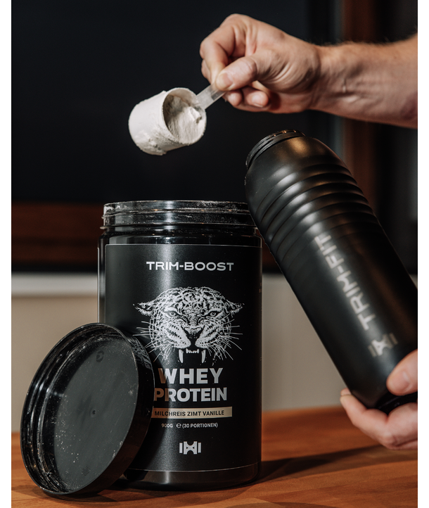 Handschaufeln von Molkenproteinpulver aus einem schwarzen Behälter mit der Aufschrift Trim-Boost Whey Protein neben einer Flasche.