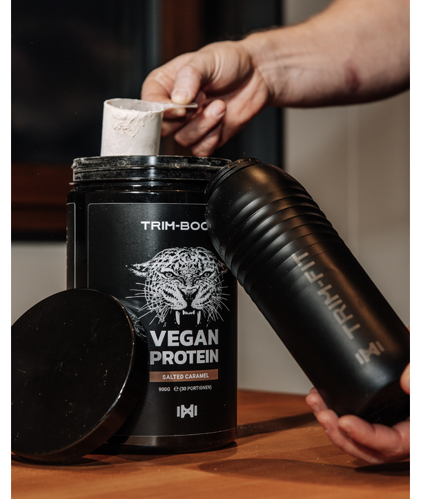 Eine Hand löffelt veganes Proteinpulver aus einem Behälter in eine schwarze Shakerflasche.