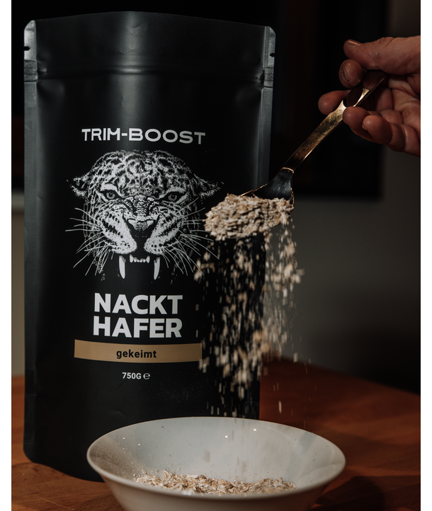 Eine Hand schüttet Haferflocken von einem Löffel in eine Schüssel, im Hintergrund ist eine Trim-Boost Nackt Hafer-Verpackung zu sehen.
