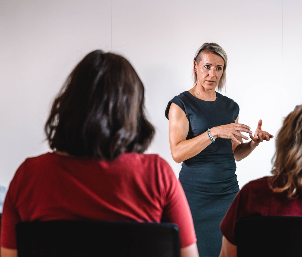 Eine Frau spricht zu zwei sitzenden Personen in einem Klassenzimmer oder in einer Besprechung.