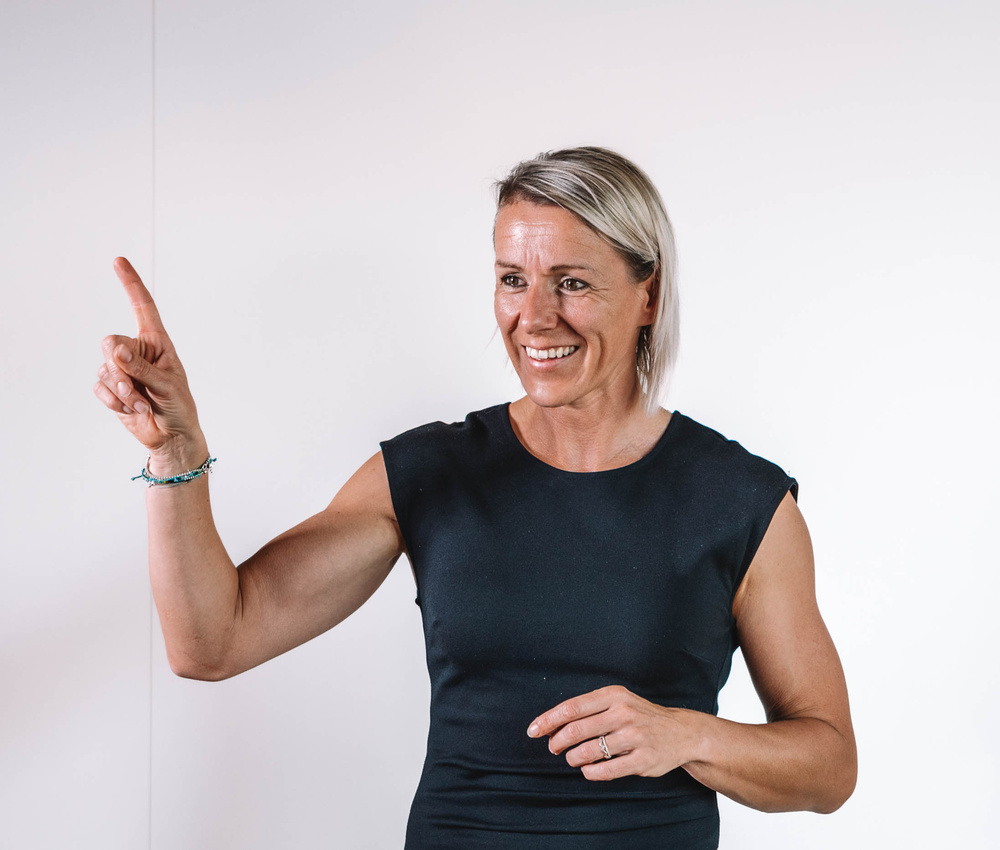 Eine lächelnde Frau in einem schwarzen Kleid zeigt mit ihrem rechten Zeigefinger vor einem schlichten weißen Hintergrund nach oben.