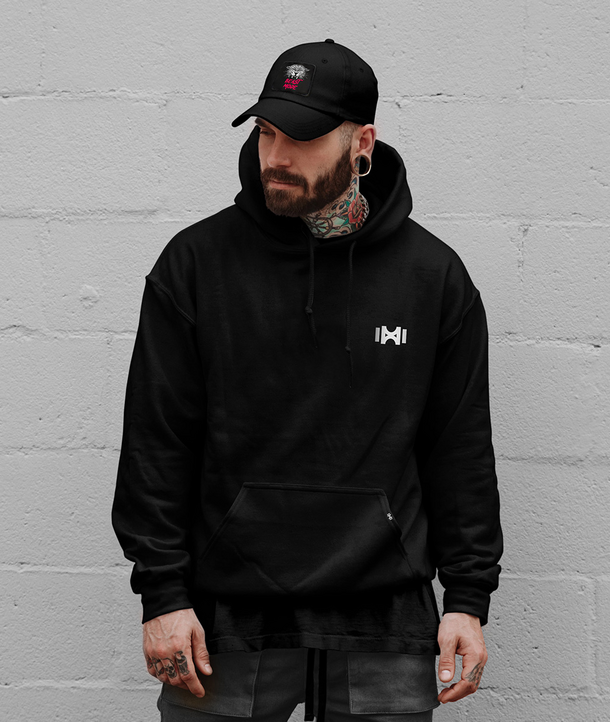 Mann in schwarzem Kapuzenpulli und Mütze mit Tätowierungen, der vor einer hellgrauen Backsteinmauer steht.