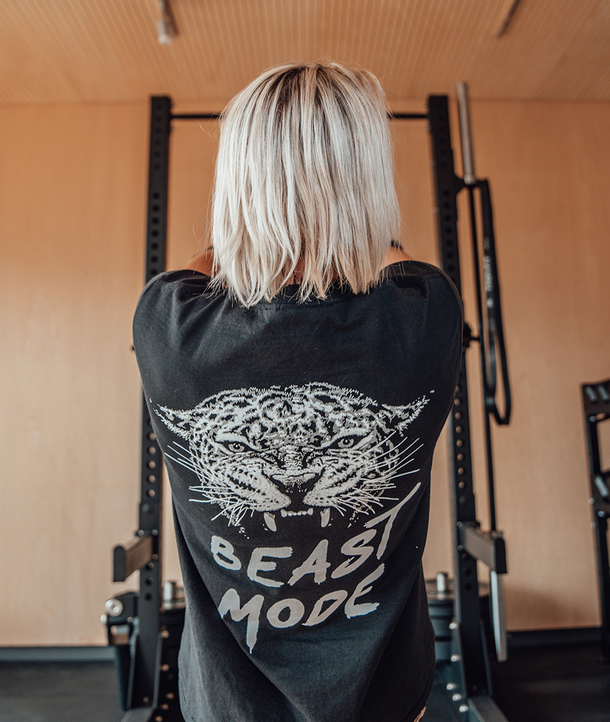 Eine Person mit blonden Haaren, die ein Beast Mode-Shirt trägt, steht vor einem Fitnessgerät und schaut weg.