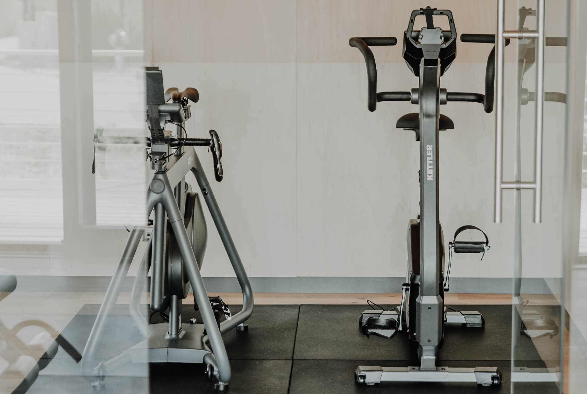 Zwei stationäre Heimtrainer stehen nebeneinander auf schwarzen Matten in einer modernen Turnhalle.