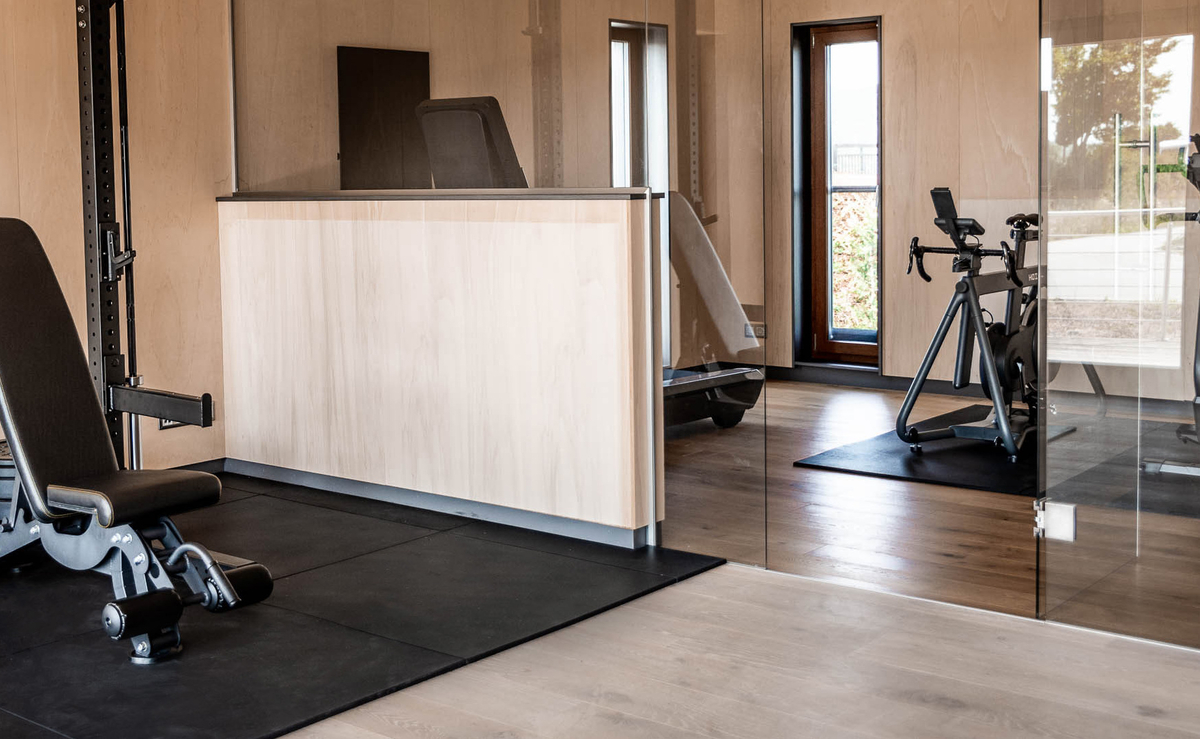 Modernes Fitnessstudio mit einer Bank, Hanteln, einem Heimtrainer und großen Fenstern für natürliches Licht.