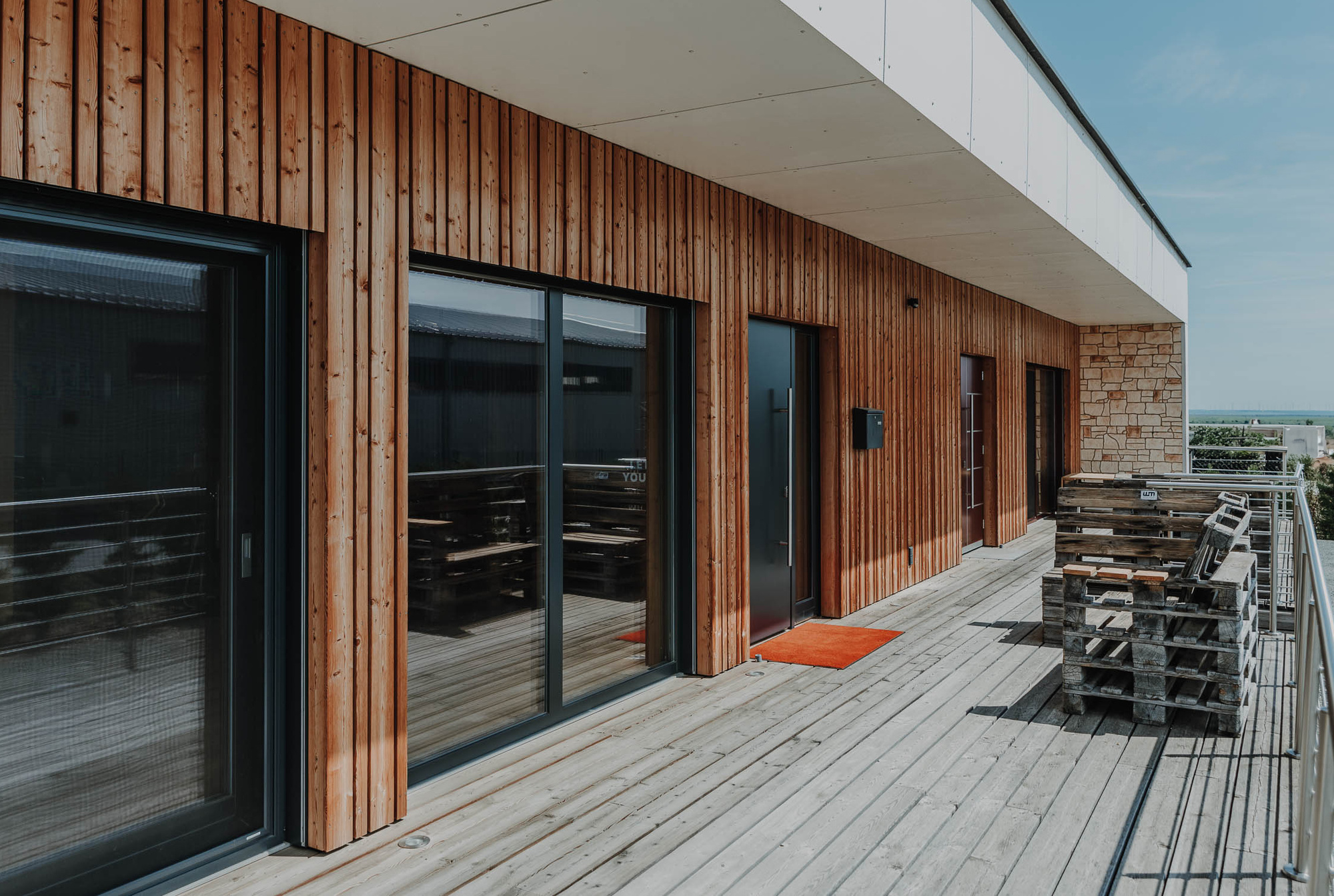 Modernes Holzgebäude mit großen Glastüren, einer Terrasse und gestapelten Holzpaletten an einem sonnigen Tag.
