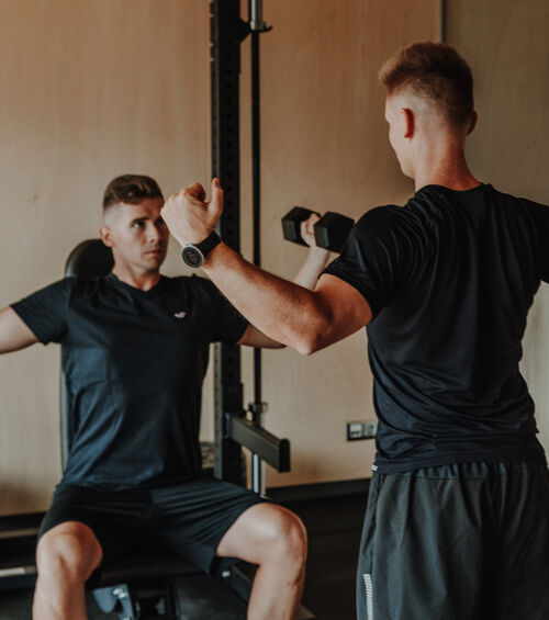 Zwei Männer trainieren in einem Fitnessstudio, einer sitzt mit erhobenen Armen, der andere steht und hält eine Hantel.