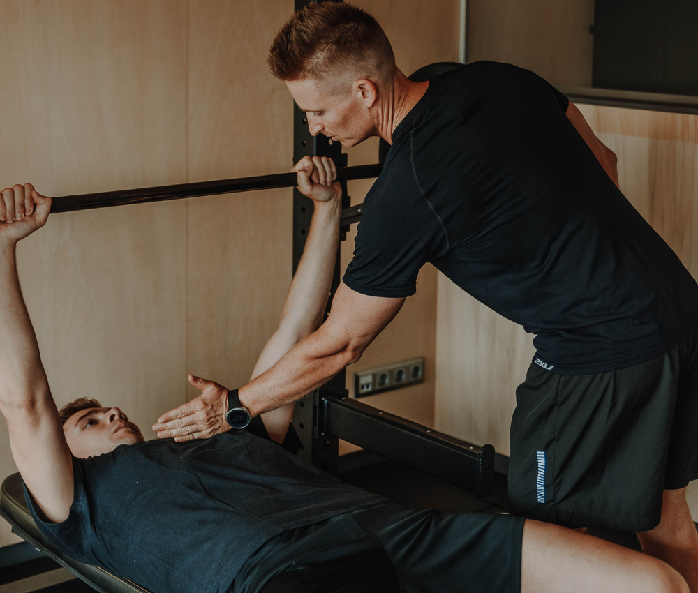 Ein Mann sieht einen anderen Mann beim Bankdrücken in einem Fitnessstudio eine Langhantel heben.