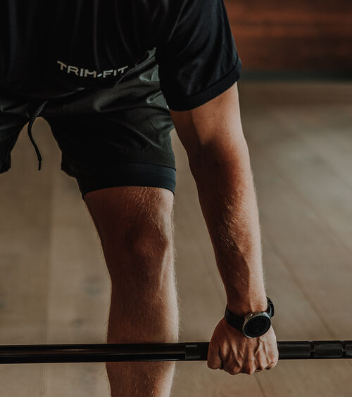 Person, die in einer Halle eine Hantel hebt und dabei schwarze Shorts, ein schwarzes T-Shirt und eine Armbanduhr trägt.