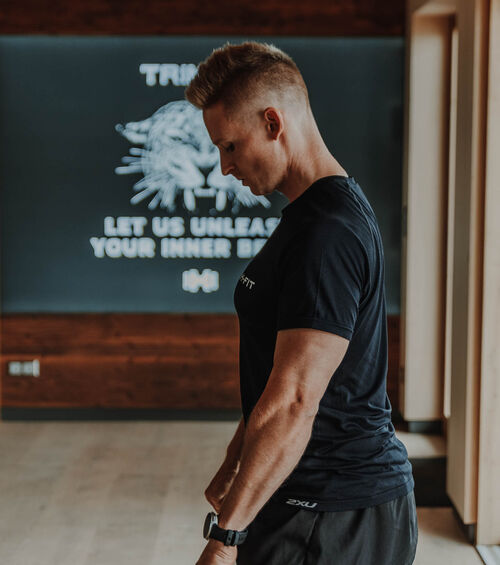 Ein Mann in Sportkleidung steht seitlich in einer Turnhalle und blickt nach unten, während im Hintergrund ein Motivationsschild zu sehen ist.