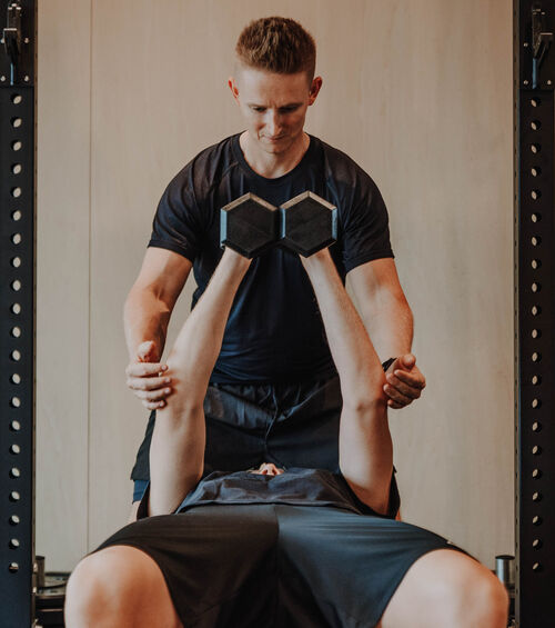 Ein Trainer sieht eine Person, die in einem Fitnessstudio auf einer Bank liegend Hanteln hebt.