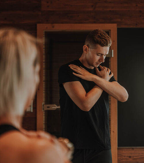 Ein Mann in einem schwarzen Hemd verschränkt die Arme vor der Brust und streckt sich vor einem Spiegel, während eine andere Person im Vordergrund verschwimmt.