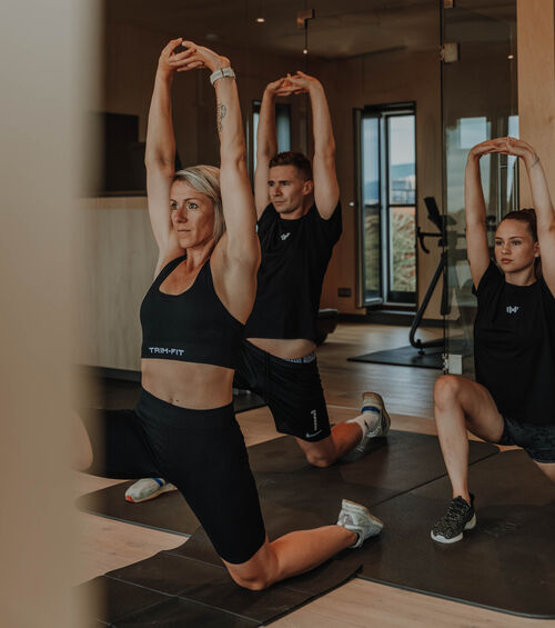Drei Personen in Sportkleidung dehnen sich mit erhobenen Armen in einer Turnhalle oder einem Fitnessstudio.