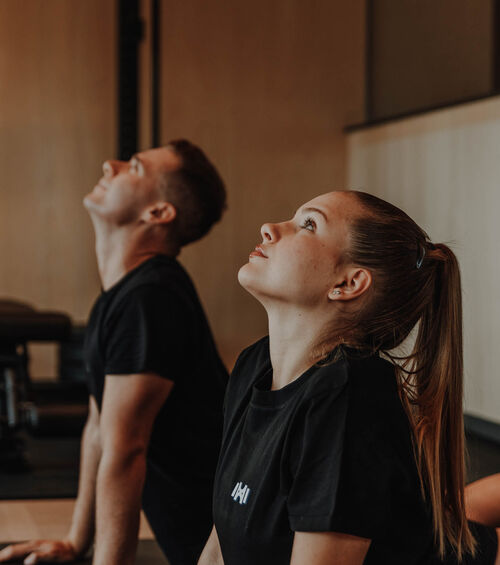 Zwei Personen in schwarzen Hemden machen eine Yoga-Dehnung und schauen in einem schwach beleuchteten Raum nach oben.
