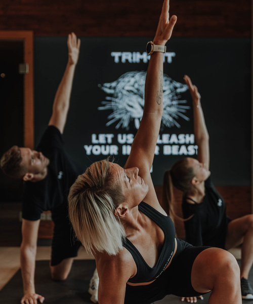Drei Personen in Sportkleidung dehnen sich mit einem erhobenen Arm in einer Turnhalle nach einem Gruppenfitnesskurs.