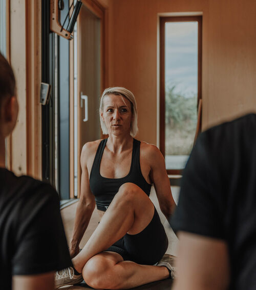 Frau in Trainingskleidung macht eine sitzende Twist-Yoga-Pose in einem Raum, mit Blick auf zwei Personen im Vordergrund.