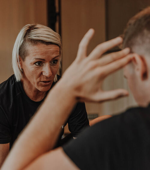 Eine Frau mit kurzen blonden Haaren hört aufmerksam einem Mann zu, der vor ihr mit der Hand gestikuliert.