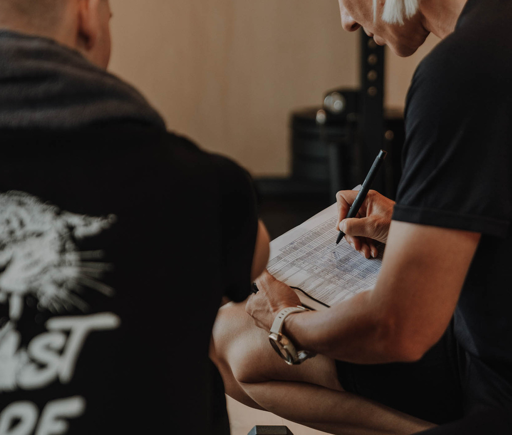 Zwei Personen sitzen, eine schreibt mit einem Stift auf einem Klemmbrett, beide tragen dunkle T-Shirts in einer Turnhalle.