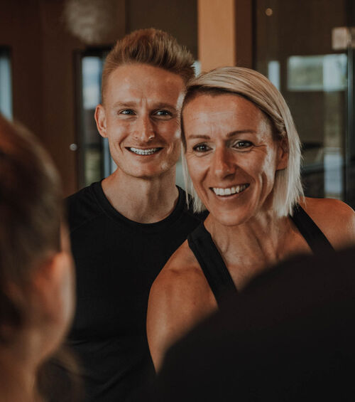 Zwei Personen, die lächeln und jemanden außerhalb des Bildes in einem Fitnessstudio ansehen.