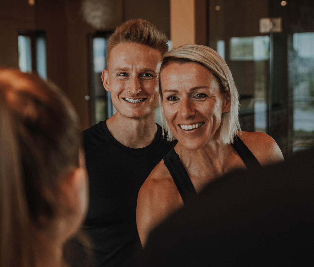 Zwei Personen, die lächeln und jemanden außerhalb des Bildes in einem Fitnessstudio ansehen.