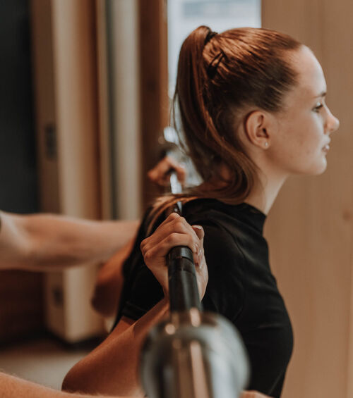 Frau mit Pferdeschwanz hebt eine Langhantel in einer Halle, unterstützt von einem Trainer.