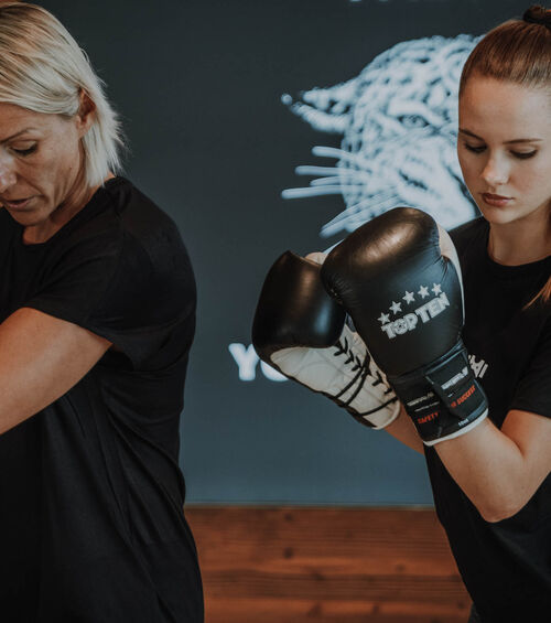 Zwei Frauen in schwarzen Hemden boxen in einer Halle, eine trägt Handschuhe, die andere steht neben ihr.