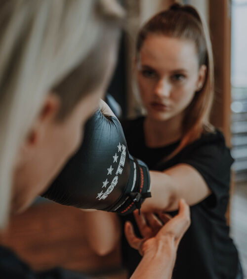 Eine Frau übt das Schlagen mit einem Boxhandschuh unter Anleitung eines Trainers, der ihren Arm hält.