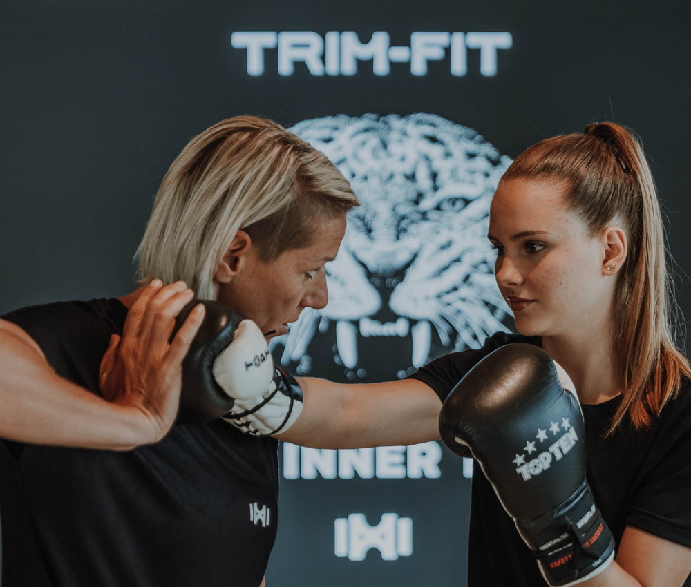 Zwei Frauen beim Boxen, von denen eine einen Schlag abwehrt, mit einem Tiger-Logo und dem Text TRIM-FIT im Hintergrund.