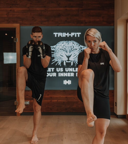 Zwei Personen in schwarzer Trainingskleidung üben Kickboxen in einem Fitnessstudio mit einem Tiger-Logo hinter ihnen.