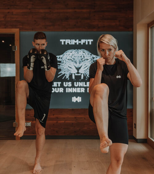 Zwei Personen in Trainingskleidung machen Kickboxing-Bewegungen in einem Fitnessstudio mit einem Trim-Fit-Schild im Hintergrund.