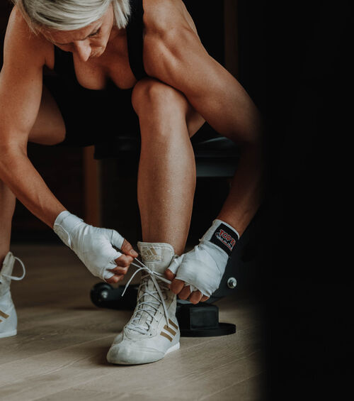 Person mit kurzen blonden Haaren, die Schnürsenkel bindet, in Trainingskleidung und mit Handschuhen, die sich auf das Training vorbereiten.