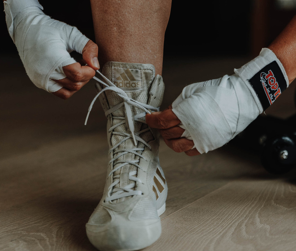 Nahaufnahme von Händen mit Boxbandagen, die die Schnürsenkel eines hellen Adidas-Boxschuhs binden.