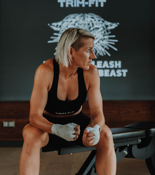 Eine sportliche Frau mit kurzen blonden Haaren sitzt auf einer Fitnessbank und trägt Trainingskleidung und Handschlaufen.