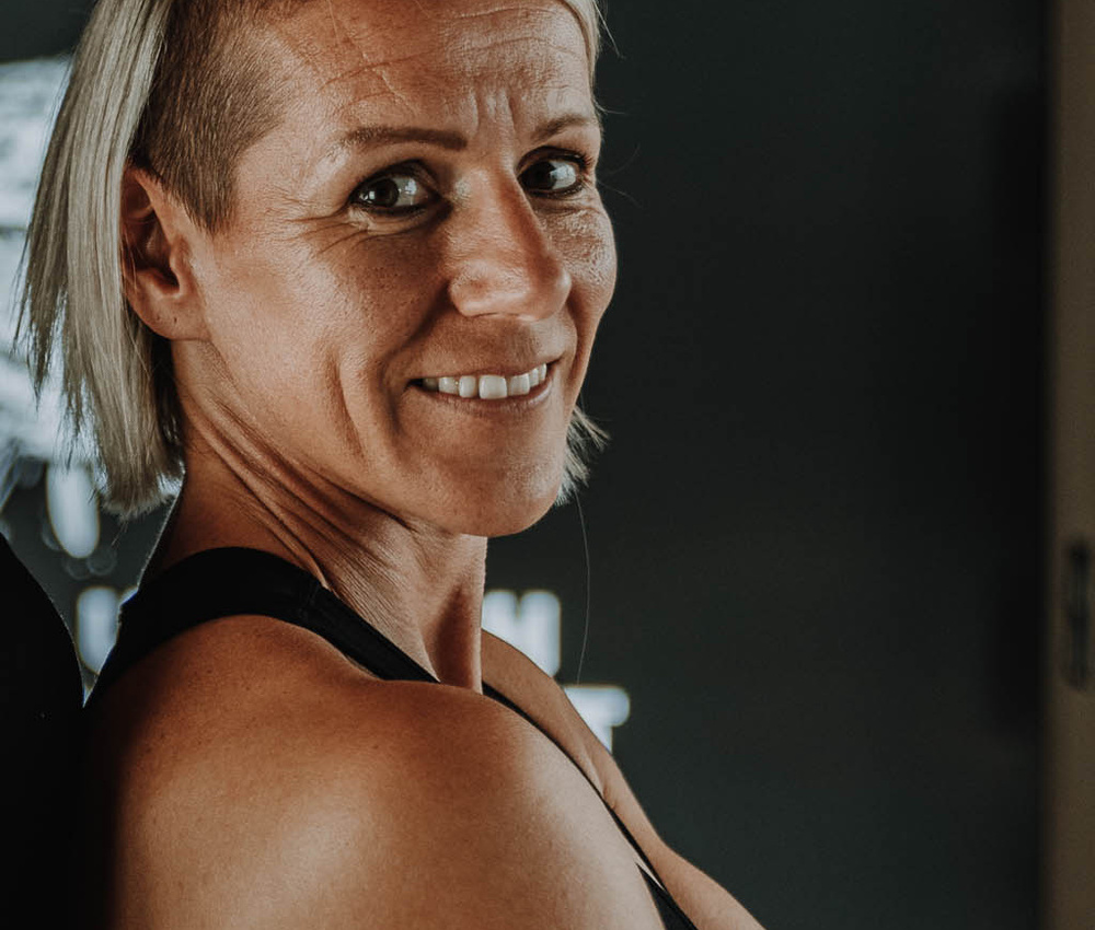 Lächelnde Person mit kurzem, blondem Haar und athletischem Körperbau, die in einer dunklen Umgebung über ihre Schulter schaut.