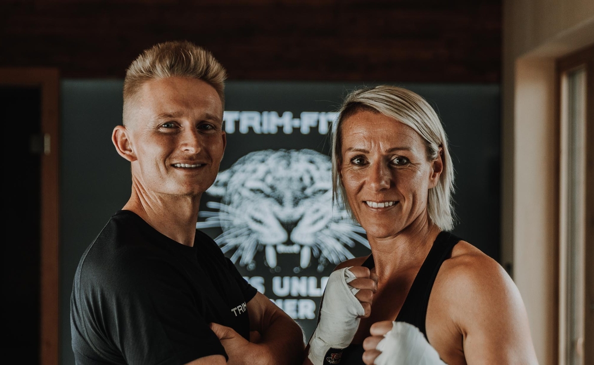 Zwei Personen in Sportkleidung lächeln und posieren mit erhobenen Fäusten in einer Turnhalle.